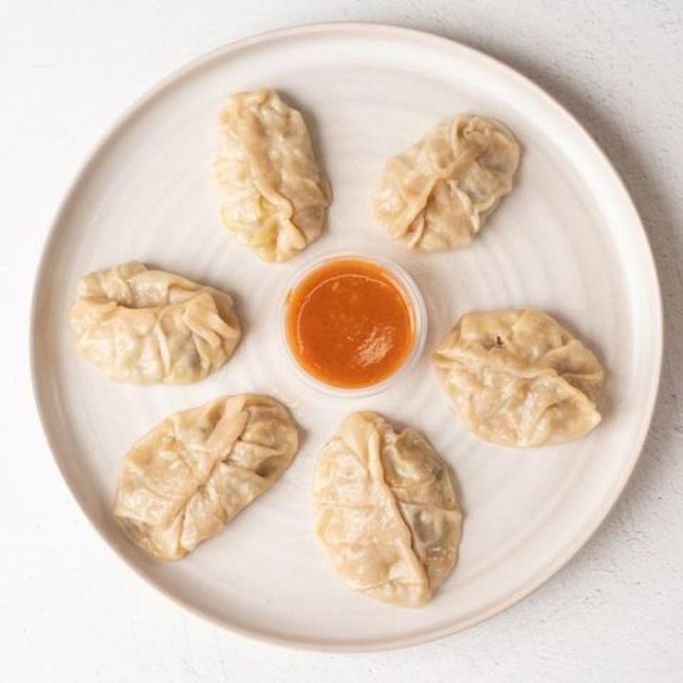 Six steamed dumplings arranged around a small cup of orange dipping sauce on a white plate.