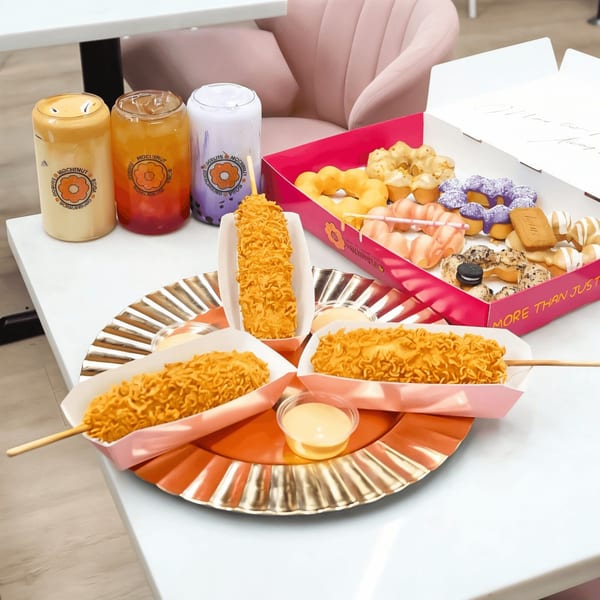 Love Me Mochi donuts, boba drinks, and Korean corn dogs with dipping sauce on a white table.