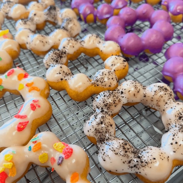 Rows of colorful Love Me Mochi donuts with various toppings on a cooling rack.