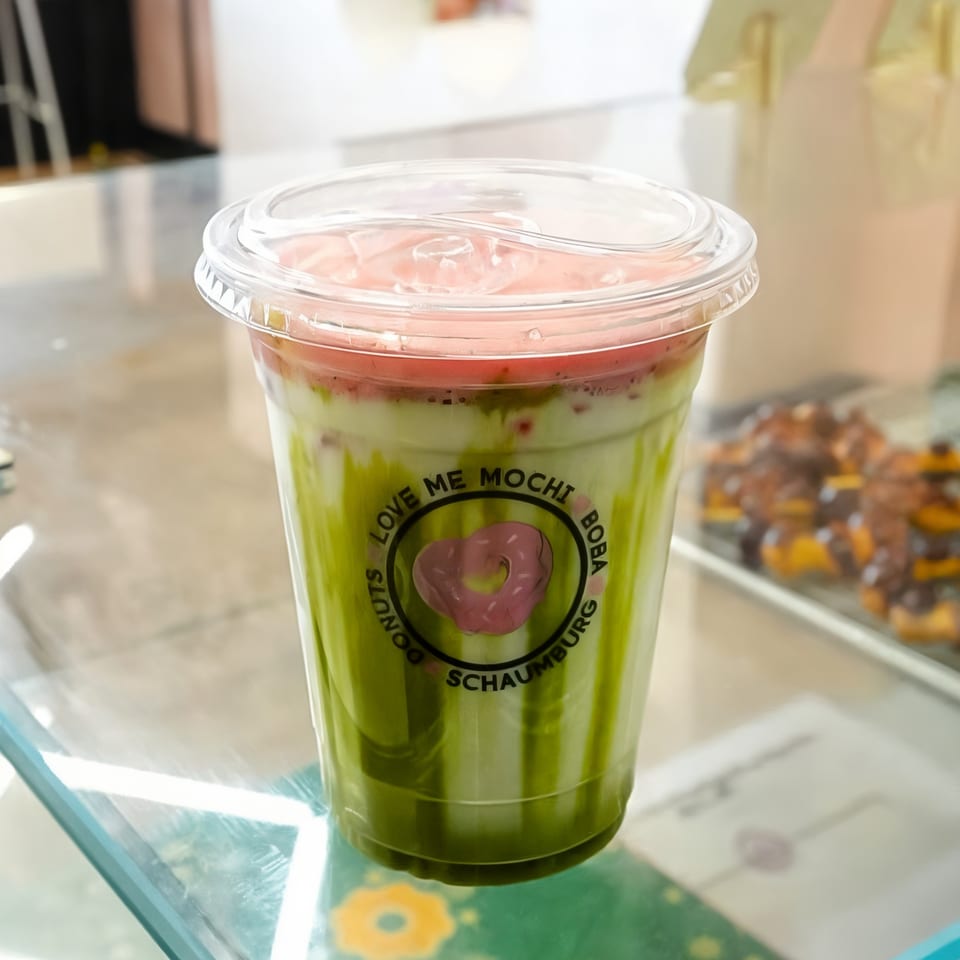 Love Me Mochi boba drink with green and pink layers, on a glass counter with donuts in background.