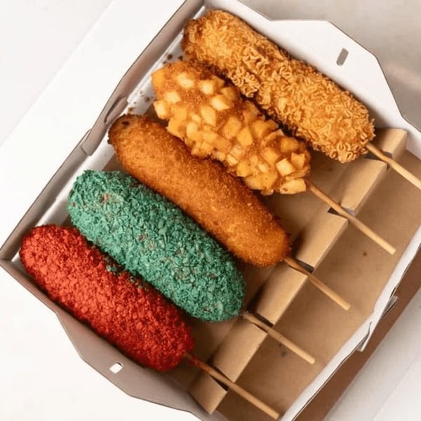Five colorful Korean corn dogs with various toppings in a white takeout box.