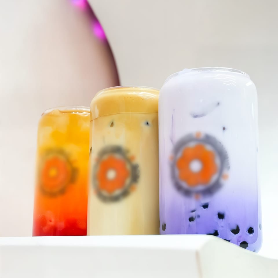 Three colorful Love Me Mochi bubble teas, orange, beige, and purple, with tapioca pearls.